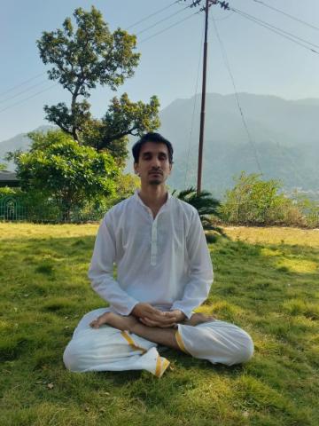 A yogi in white traditional clothing meditating outdoors with Himalaya behind him